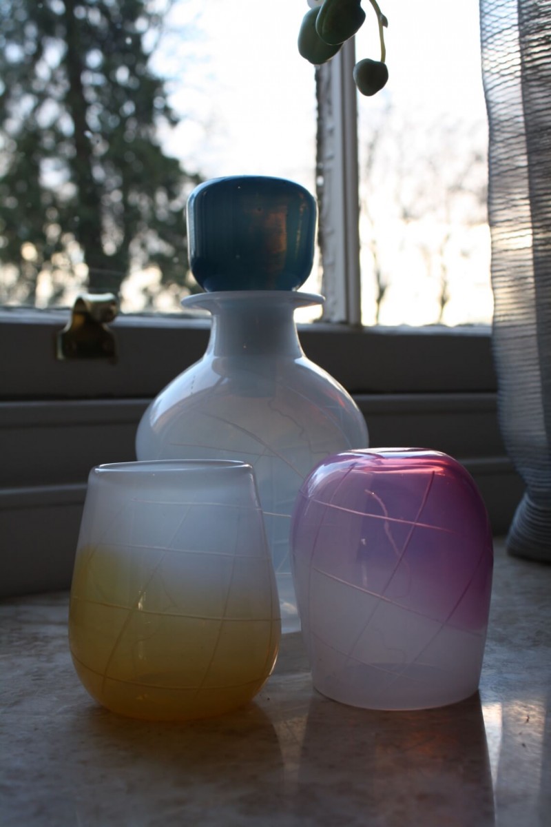 Glass decanter and tumblers in front of a window. The decanter is white with a blue stopper and the glasses are white with blue and yellow.